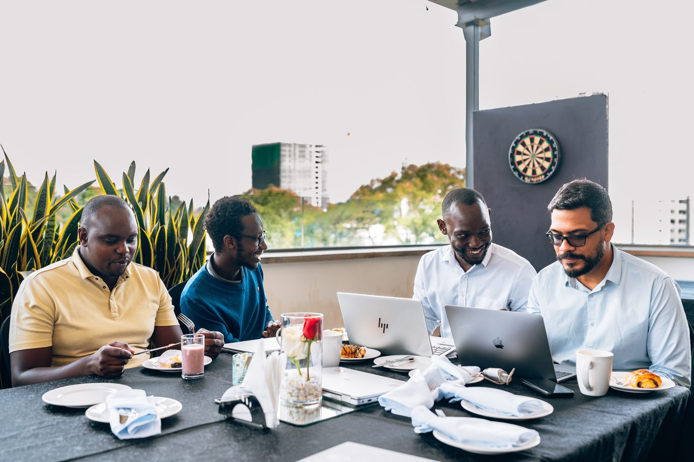 Four men at a long breakfast table with laptops, daylight, planters in background