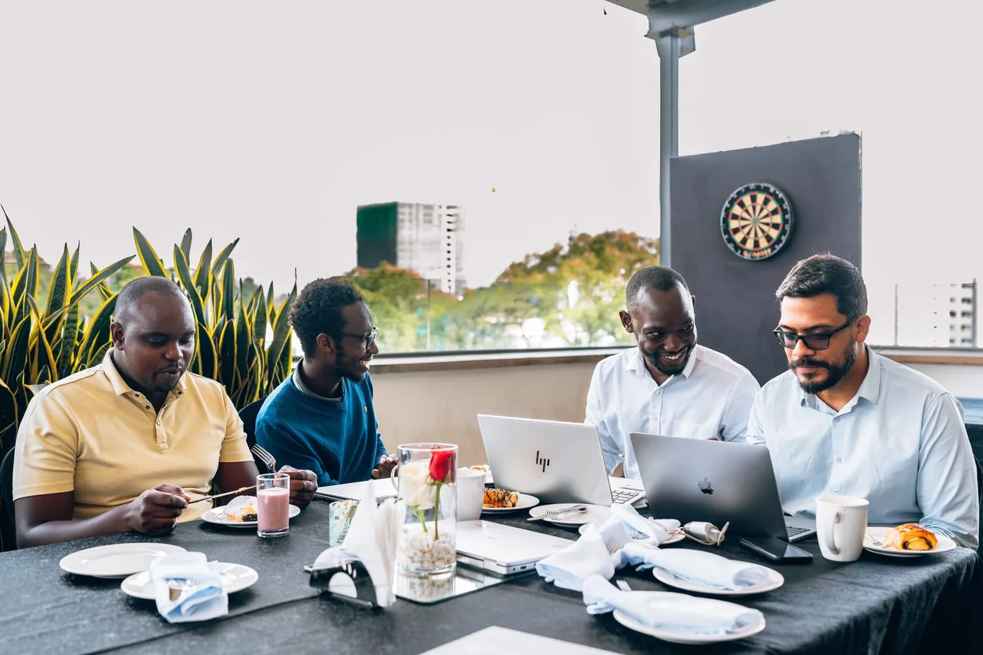 Four men at a long breakfast table with laptops, daylight, planters in background