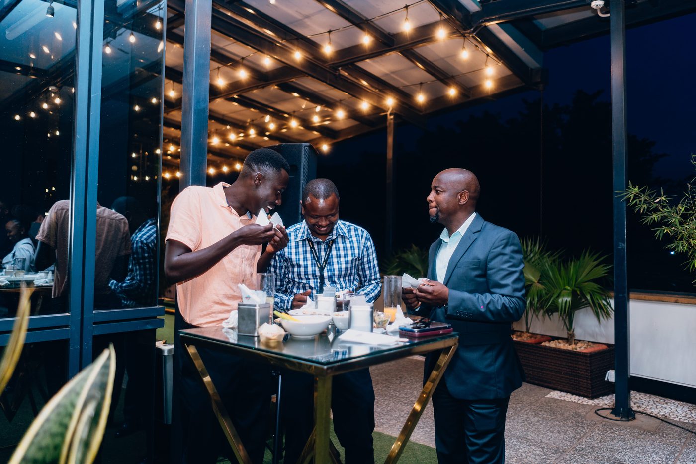 Three men in candid conversation at a low-lit corner table, sharing food and a moment