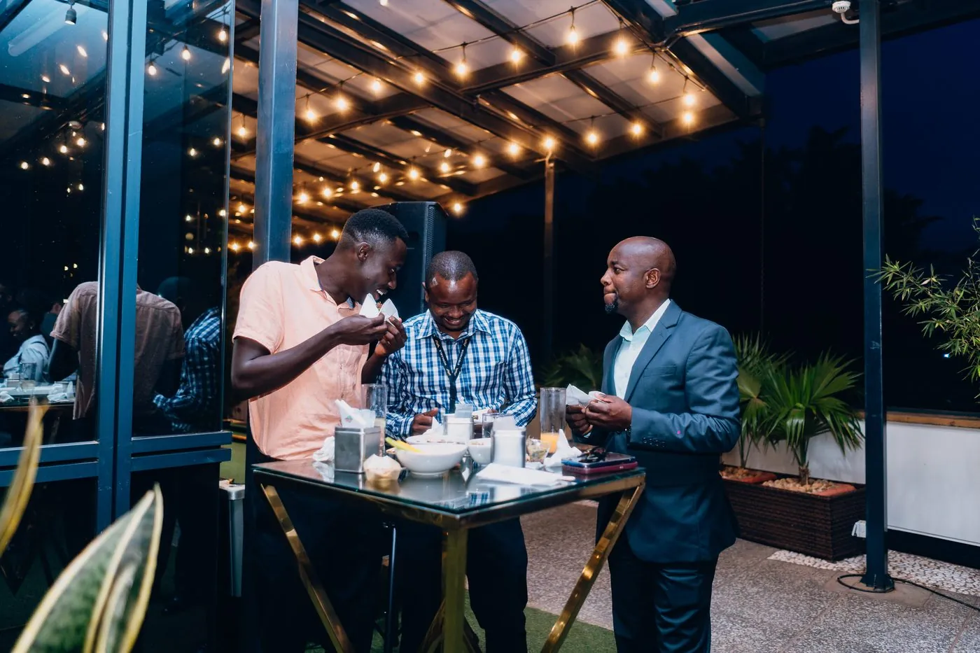 Three men in candid conversation at a low-lit corner table, sharing food and a moment