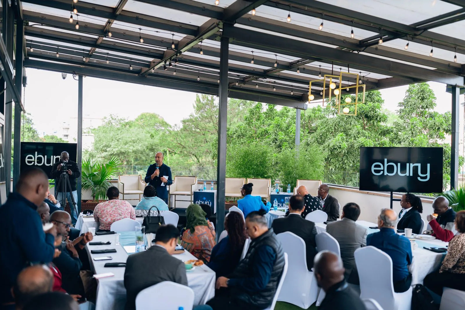 Wide view of the rooftop breakfast audience seated at white tables with Ebury signage on either side