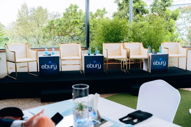 Empty panel stage with Ebury-branded cubes and cream chairs against greenery