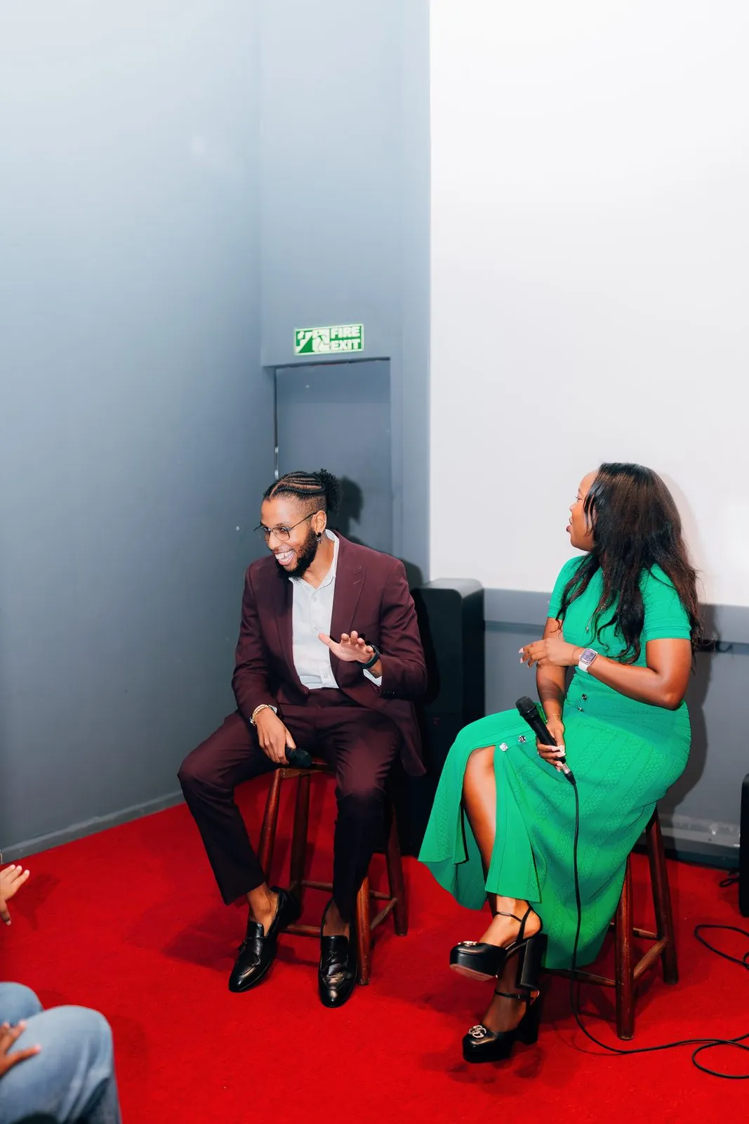 Two speakers on stools on a red carpet in front of the screen