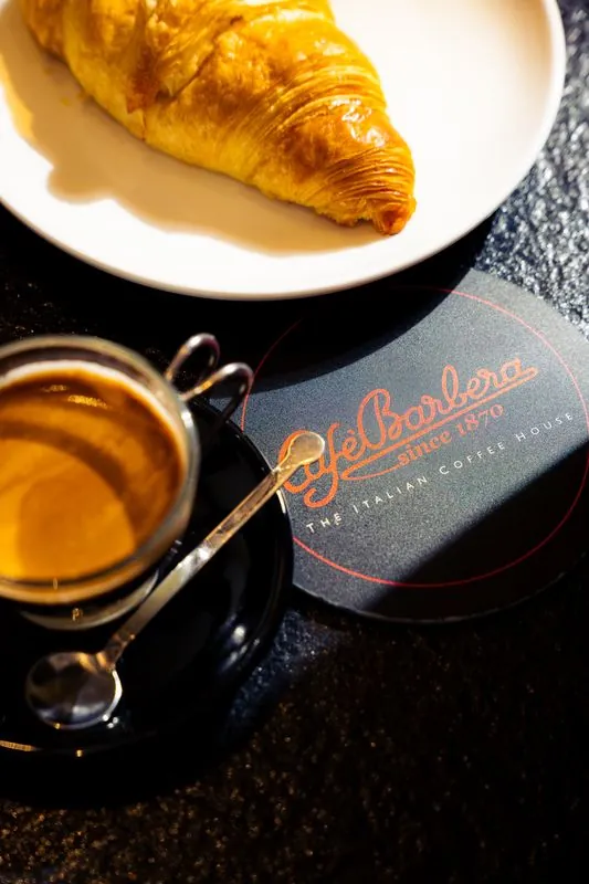 Espresso in a glass cup beside a croissant on a dark table