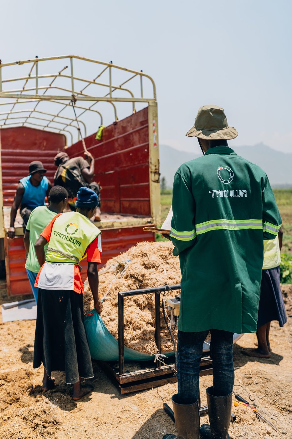 Workers loading a red truck with biomass while a supervisor in a green Tamuwa coat checks a hanging scale