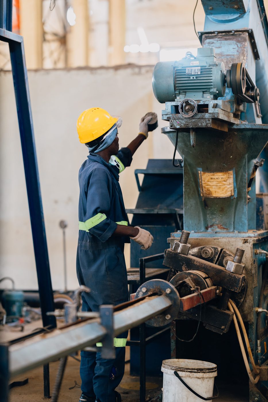 Worker in navy coveralls and yellow hard hat servicing a tall blue milling machine inside the plant